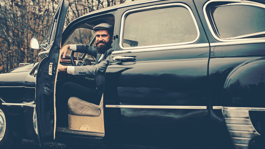 Call boy in vintage auto. Bearded man in car. Retro collection car and auto repair by mechanic driver. Travel and business trip or hitch hiking. Escort man or security guard.